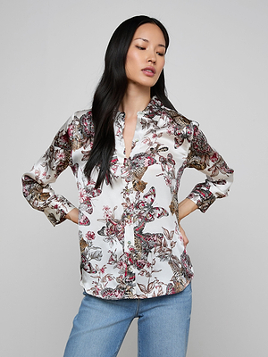 A woman with long dark hair wears the Dani Silk Blouse in a butterfly jungle print with long sleeves, paired with light blue jeans, standing against a plain light gray background.