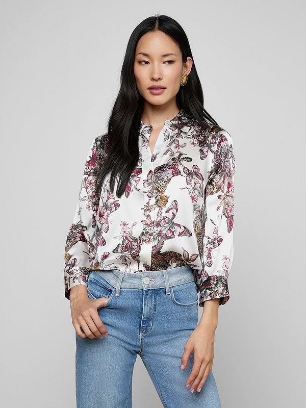 A woman wearing the Dani Silk Blouse—a patterned white top featuring floral, bird, and butterfly jungle print designs—paired with blue jeans stands against a plain background, looking slightly to the side with one hand in her pocket.