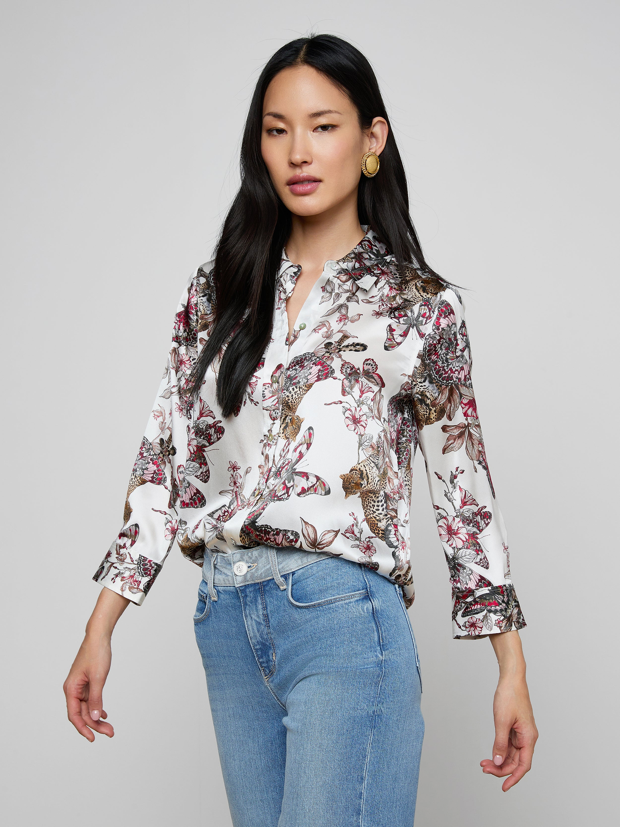 The woman is wearing the Dani Silk Blouse—a white silk button-down featuring a colorful butterfly jungle print—paired with light blue jeans. She has long dark hair and is posing against a plain light background.