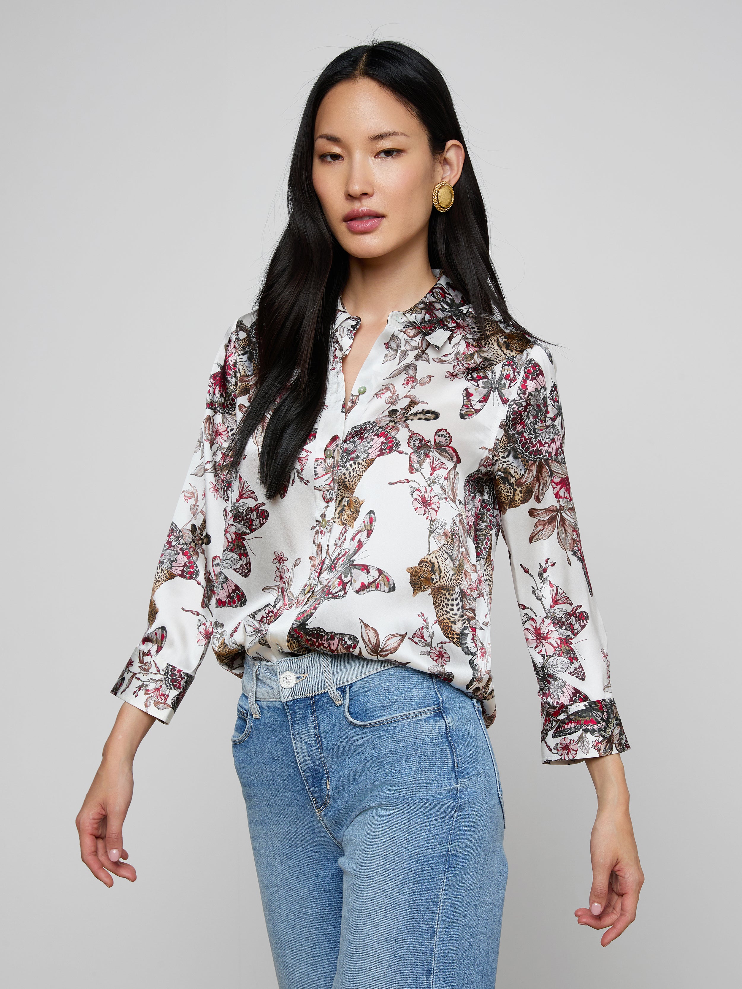 The woman is wearing the Dani Silk Blouse—a white silk button-down featuring a colorful butterfly jungle print—paired with light blue jeans. She has long dark hair and is posing against a plain light background.