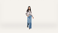A woman stands against a plain white background, wearing a floral long-sleeve blouse and wide-leg blue jeans, with one hand in her pocket and a neutral expression.