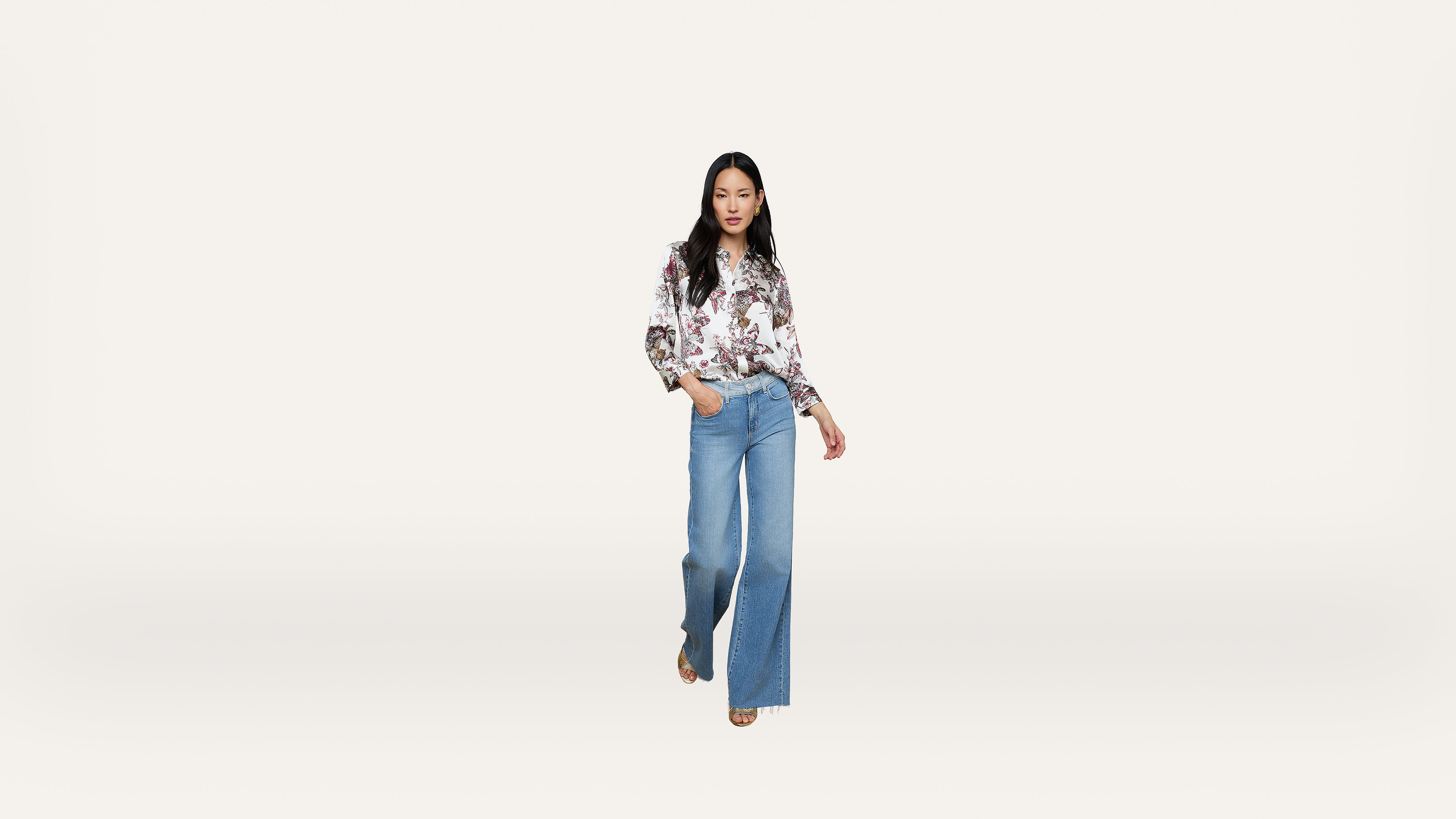 A woman stands against a plain white background, wearing a floral long-sleeve blouse and wide-leg blue jeans, with one hand in her pocket and a neutral expression.