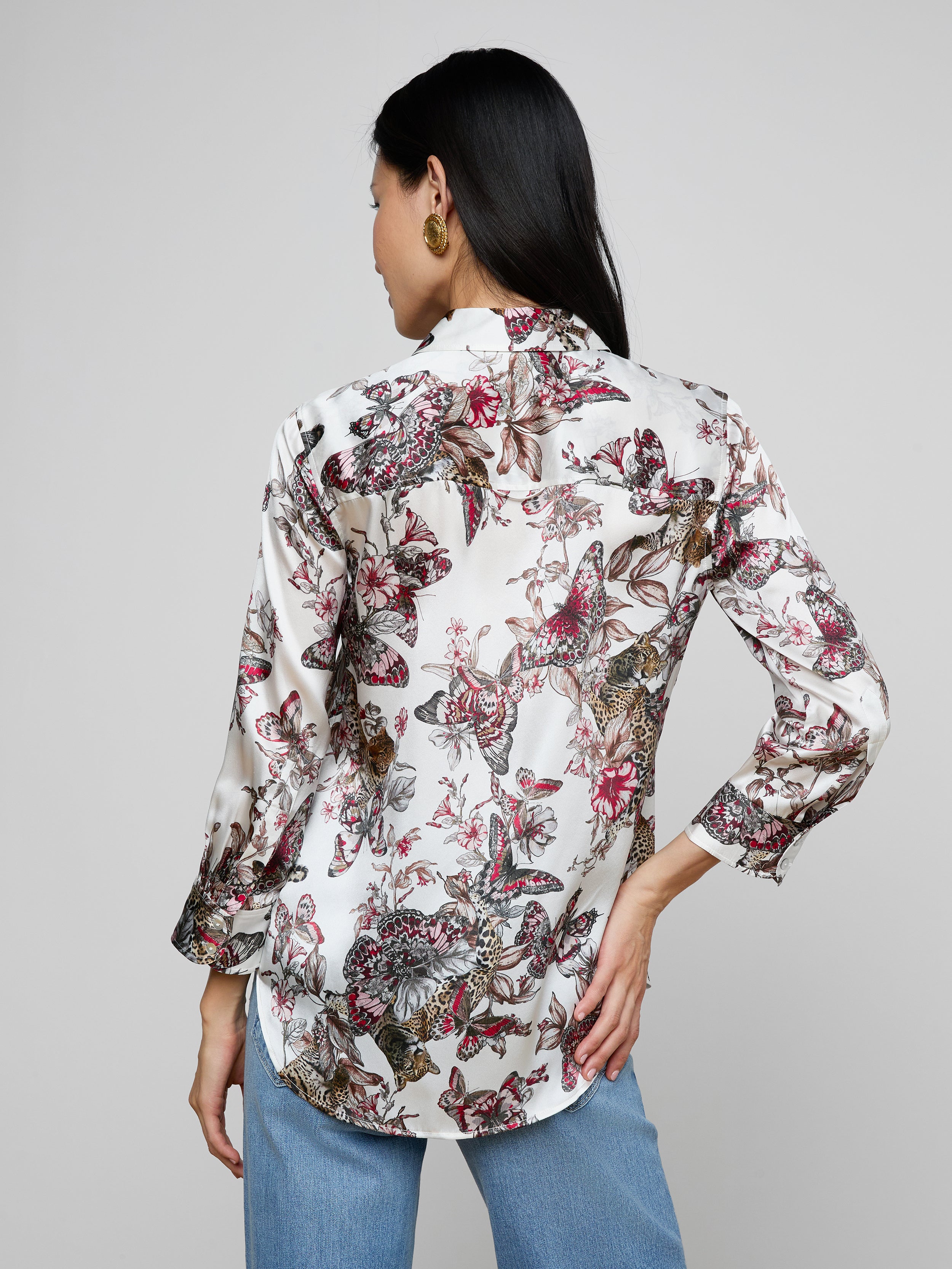 A woman is shown from the back wearing a white Dani Silk Blouse with a colorful butterfly jungle print and blue jeans, standing against a plain light background.