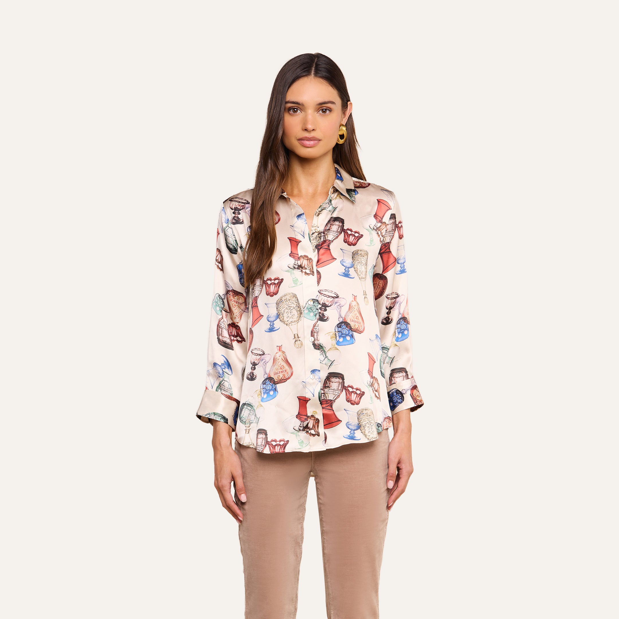 A woman with long brown hair stands against a plain background, wearing the Dani Silk Blouse featuring a light-colored base with a vibrant owl print and paired with beige pants. She looks directly at the camera with a neutral expression.