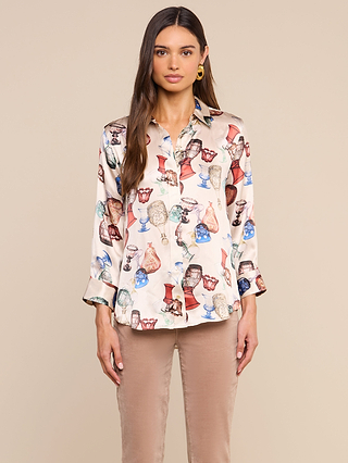 A woman stands against a plain beige background, facing forward with her hands relaxed at her sides. She wears the Dani Silk Blouse—a long-sleeve, silk button-down adorned with a vibrant vintage glassware print showcasing vases and goblets—paired with beige pants.