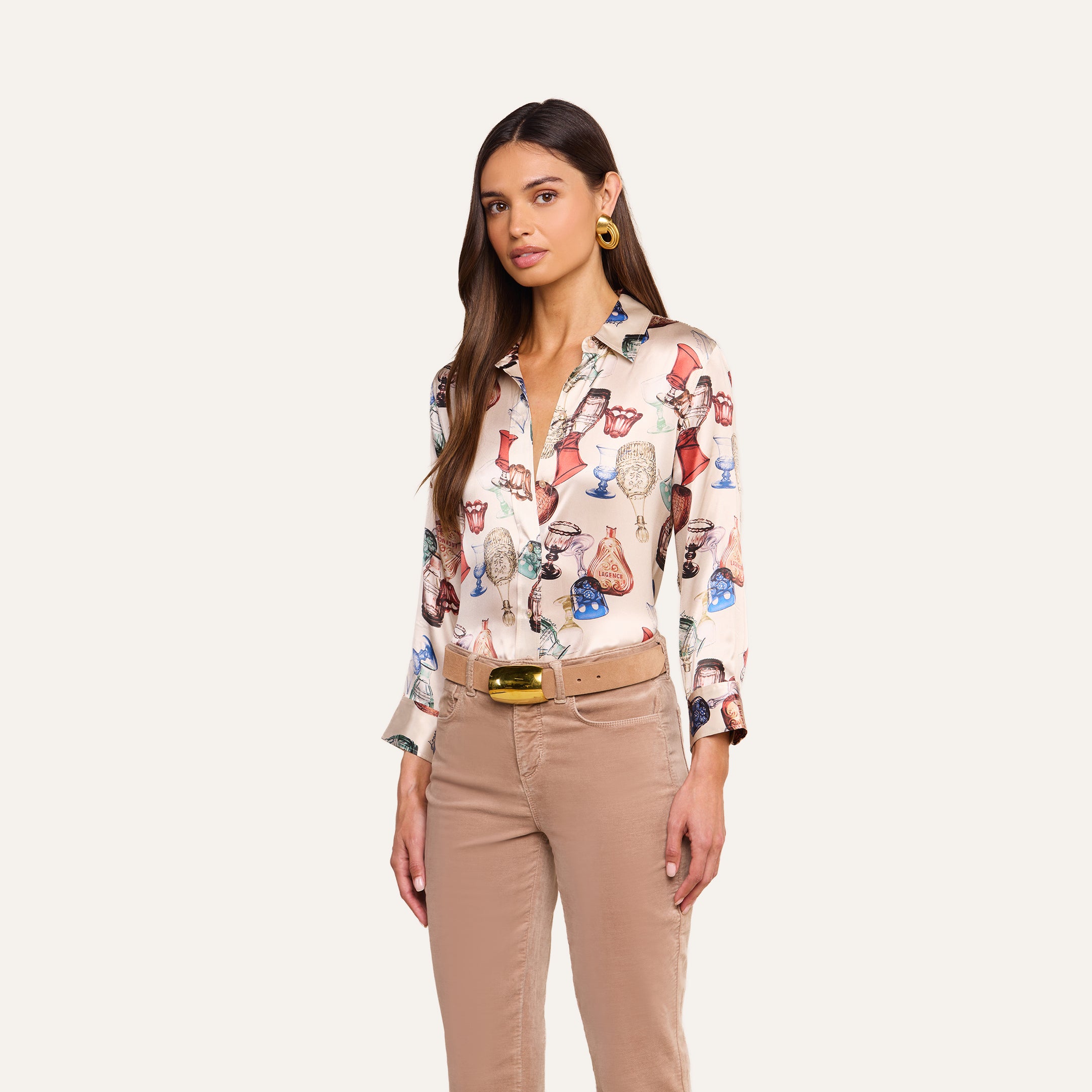 A woman with long brown hair wears the Dani Silk Blouse—a silk button-down adorned with a vintage glassware print of colorful handbags—paired with beige pants, a gold belt, and gold hoop earrings, set against a plain light background.