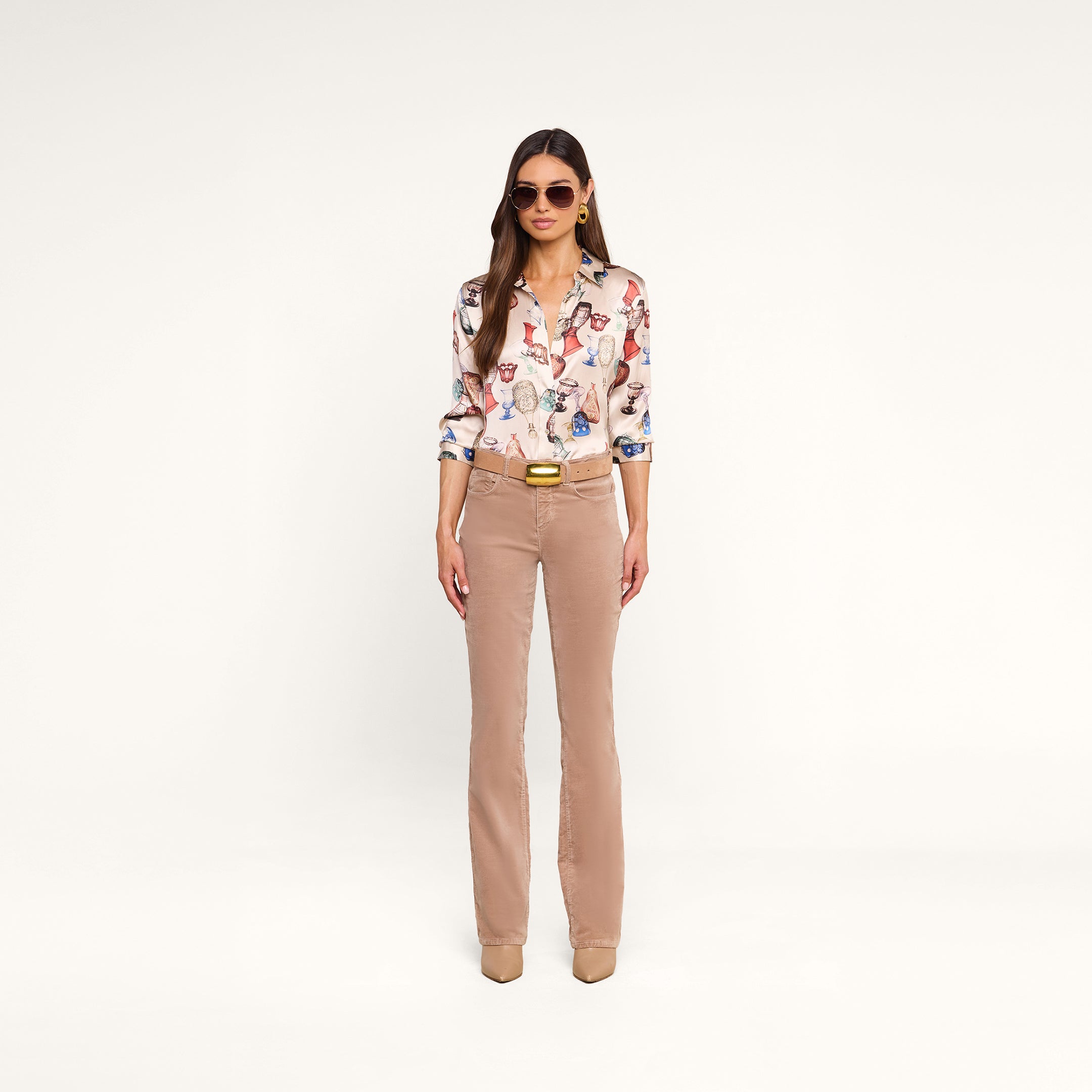 A woman wearing sunglasses, a Dani Silk Blouse featuring a vintage glassware print, beige straight-leg pants, a belt, and beige shoes stands against a plain white background.