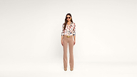 Woman standing against a plain white background, wearing sunglasses, a floral blouse, light brown flared pants, and beige shoes. She has long hair and her hands are relaxed by her sides.
