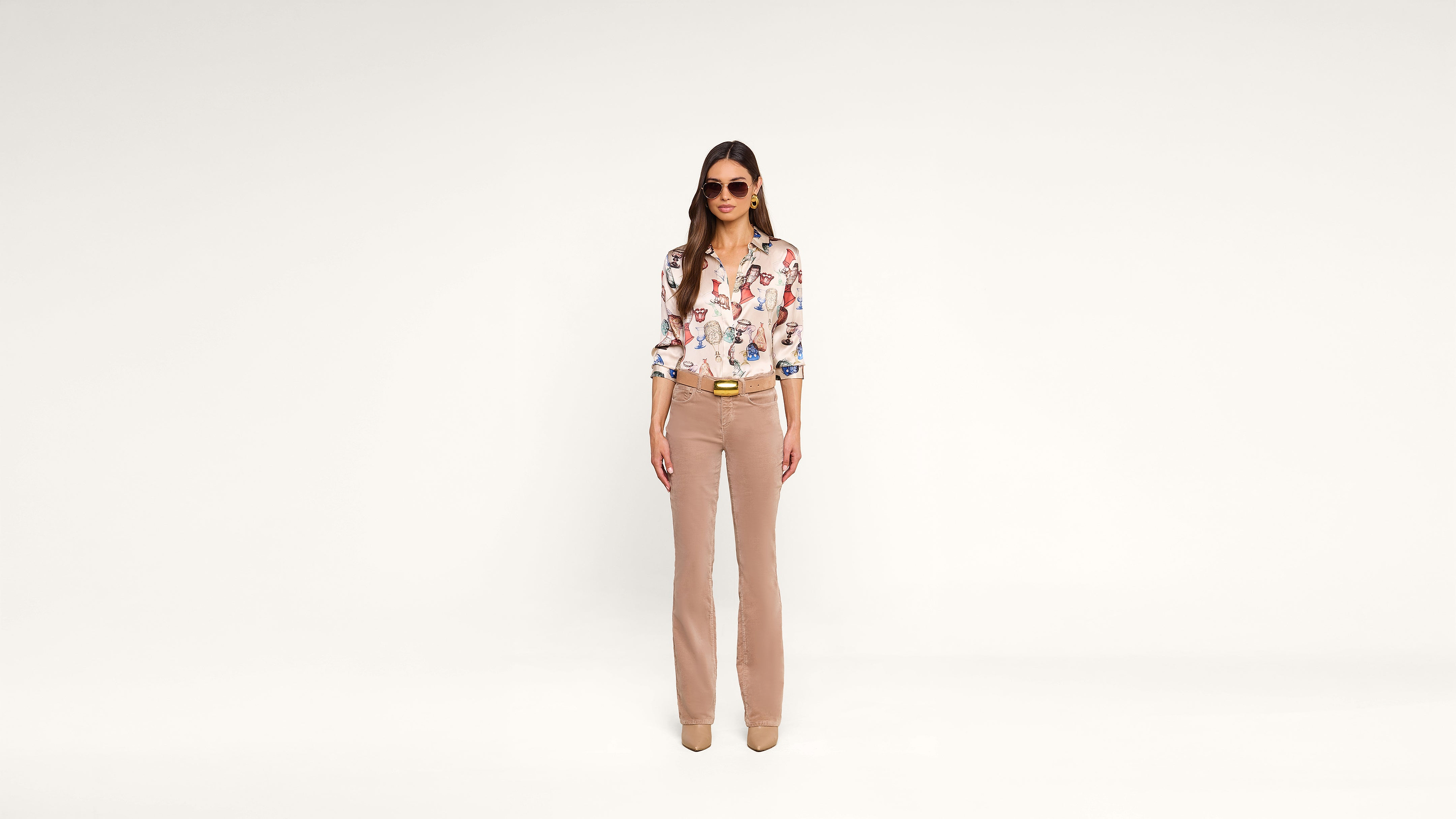 Woman standing against a plain white background, wearing sunglasses, a floral blouse, light brown flared pants, and beige shoes. She has long hair and her hands are relaxed by her sides.