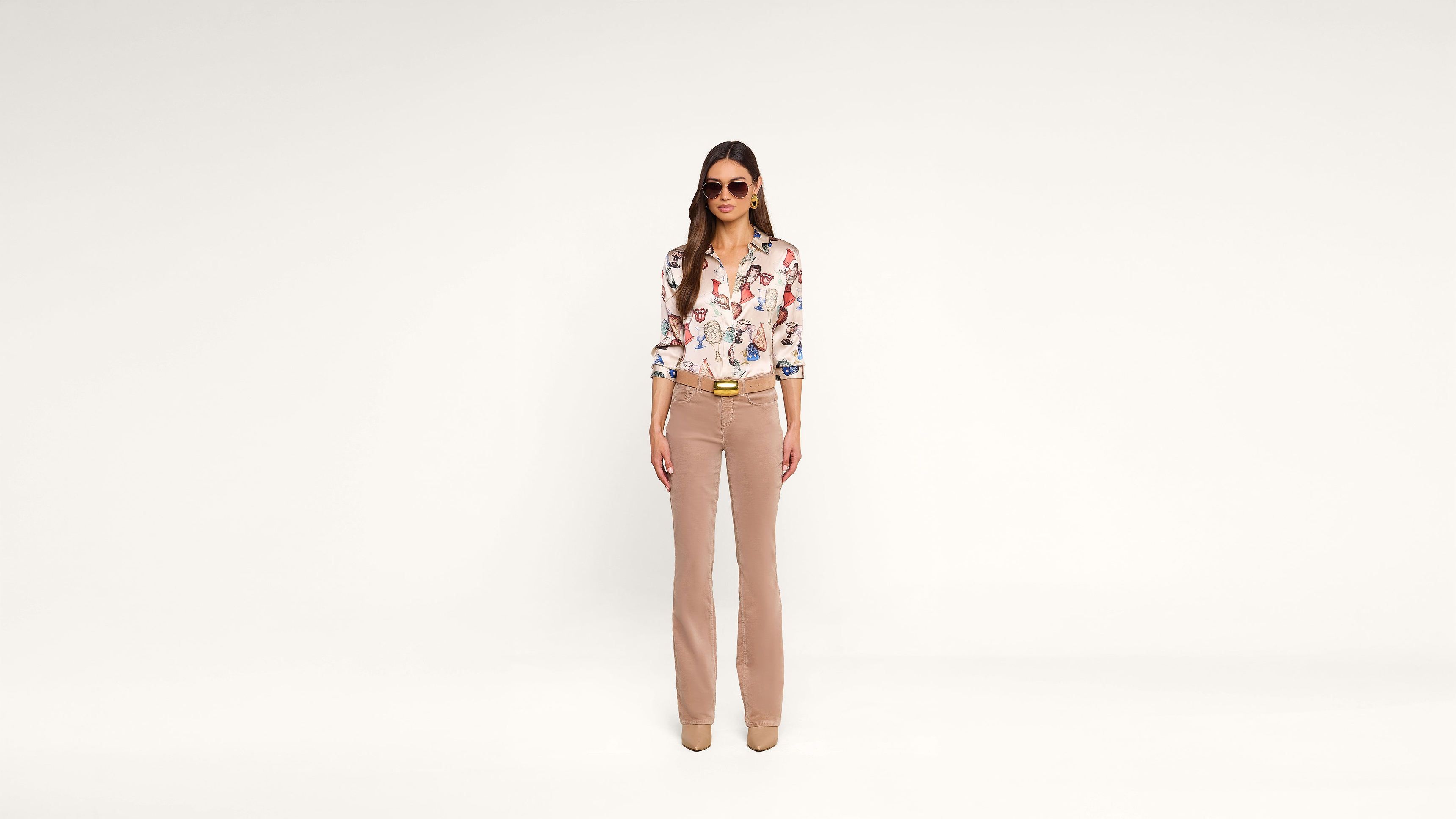 Woman standing against a plain white background, wearing sunglasses, a floral blouse, light brown flared pants, and beige shoes. She has long hair and her hands are relaxed by her sides.