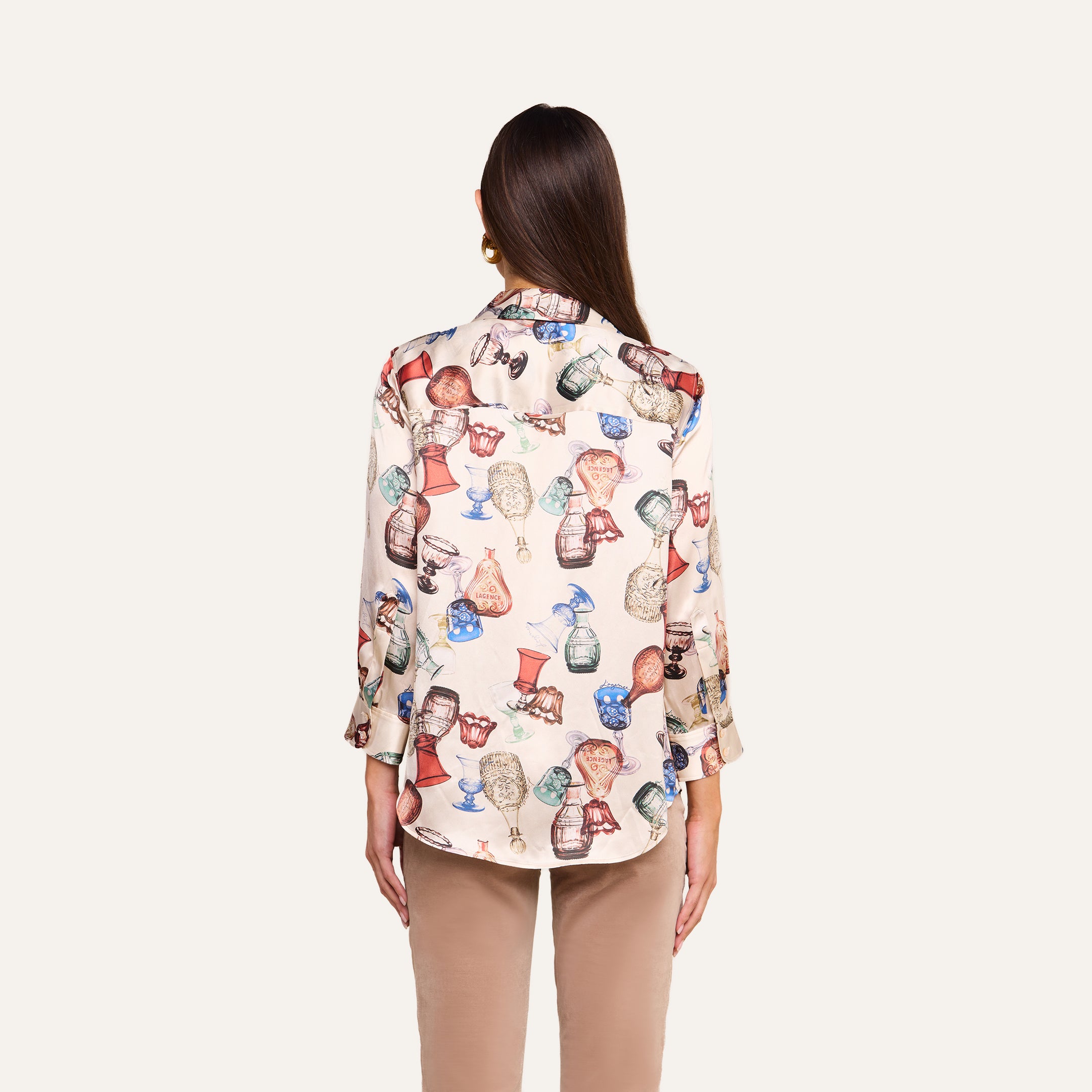 A person stands facing away, wearing a Dani Silk Blouse featuring a vintage glassware print and light brown pants against a plain, light background.