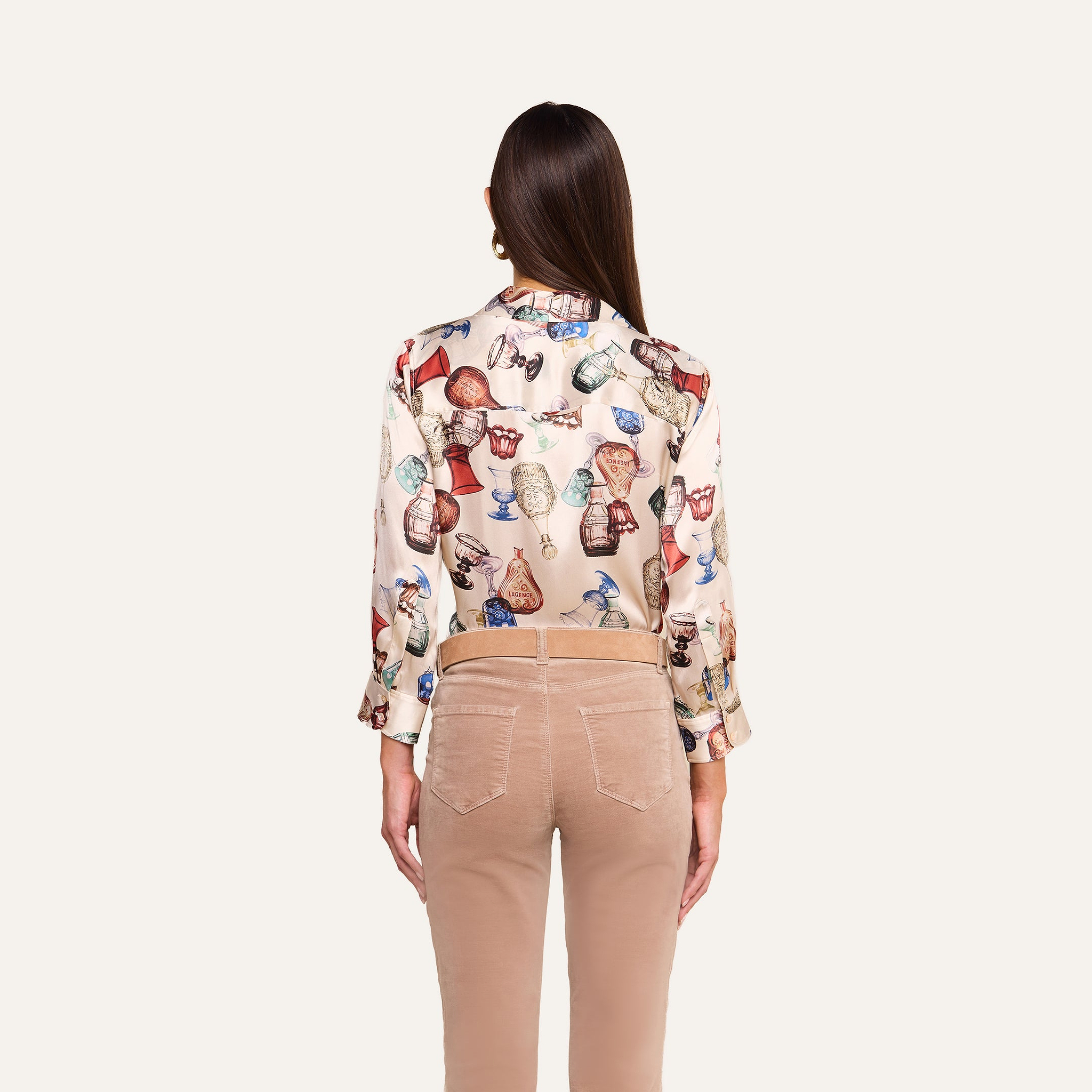 A woman stands with her back to the camera, wearing beige pants and the Dani Silk Blouse in cream, which features a vintage glassware print and pottery-inspired motifs. The background is plain white.
