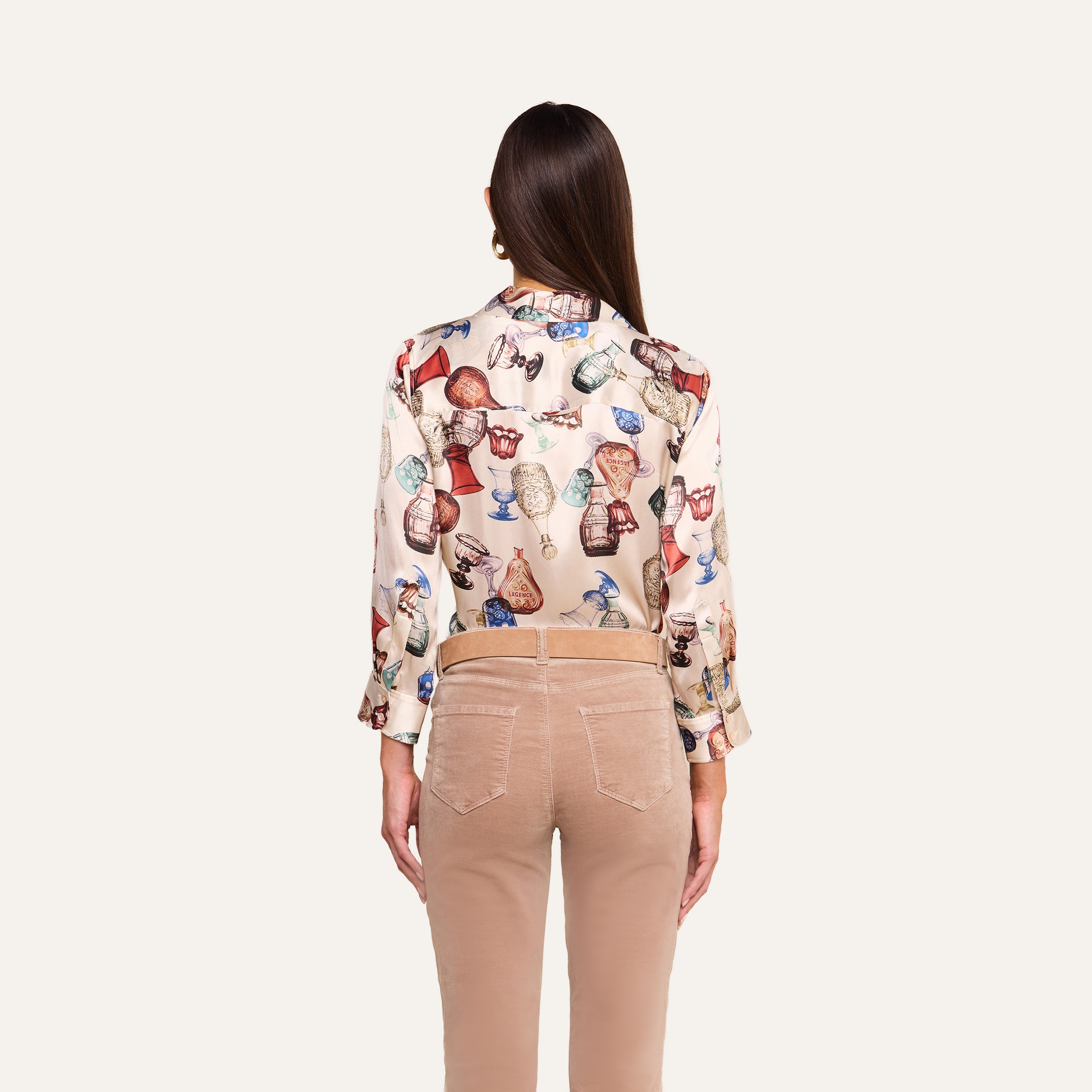 A woman stands with her back to the camera, wearing beige pants and the Dani Silk Blouse in cream, which features a vintage glassware print and pottery-inspired motifs. The background is plain white.