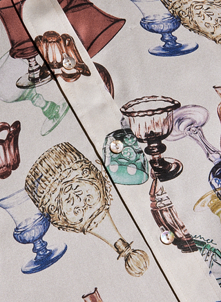 A close-up of the Dani Silk Blouse showcases its detailed, colorful illustrations of various vintage glassware—goblets and cups—printed across the fabric, complemented by white buttons along the shirt’s placket.