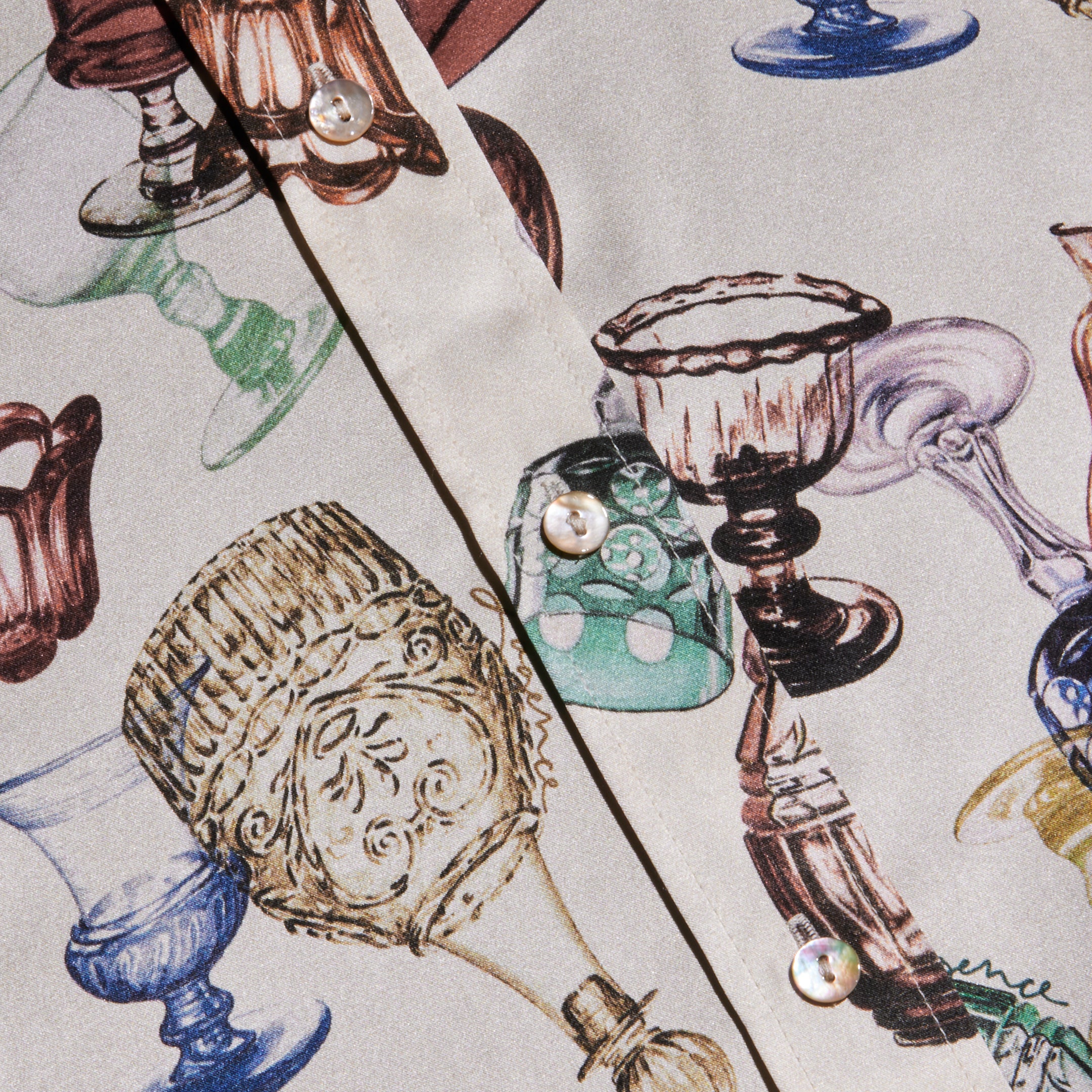 A close-up of the Dani Silk Blouse highlights a vibrant pattern of vintage glassware print goblets and cups, with pearly buttons adorning the front.