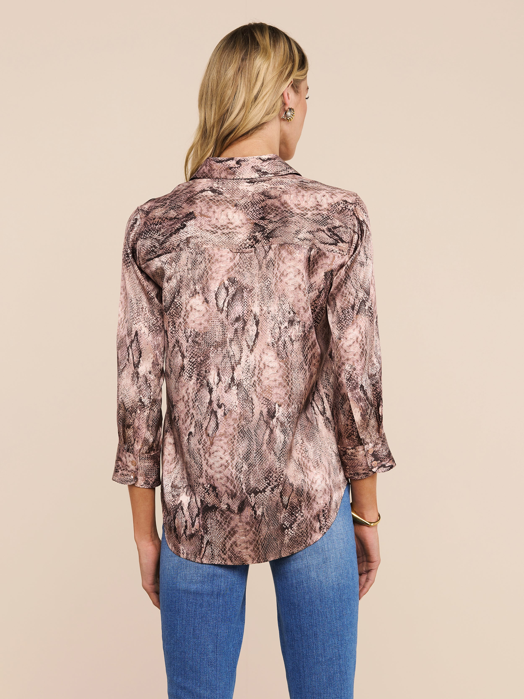 A person with long blonde hair is shown from the back against a plain, light background, wearing the Dani Silk Blouse—a snake print top with three-quarter sleeves—paired with blue jeans.