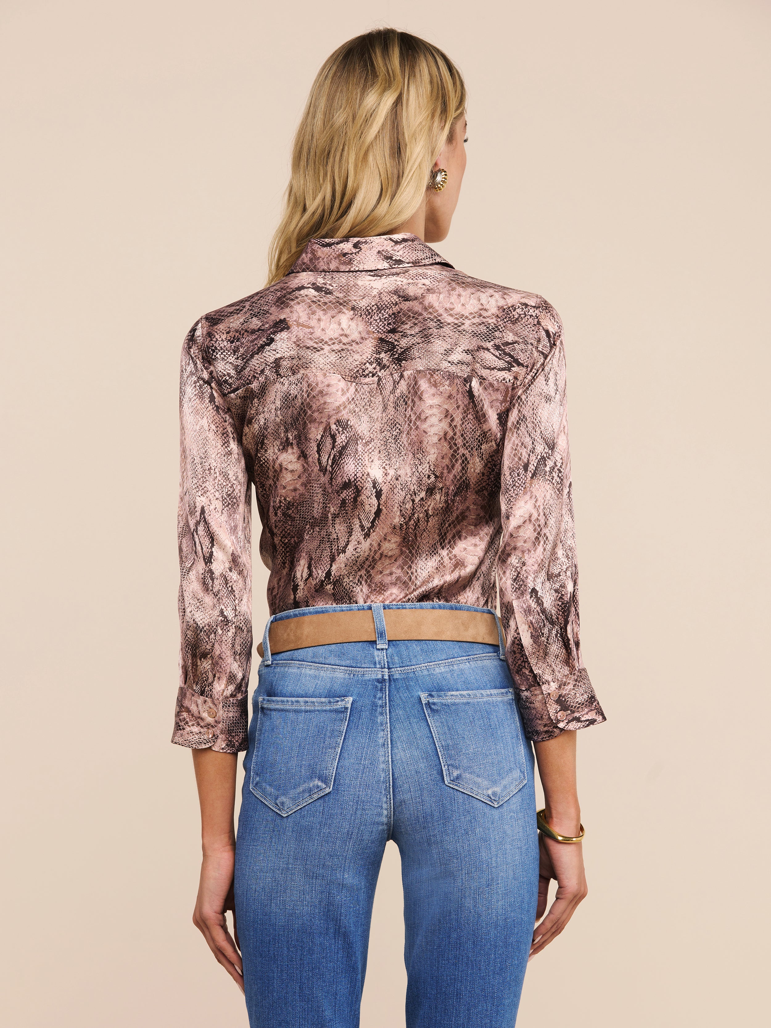 A woman with long blonde hair wears the Dani Silk Blouse, featuring a snake print and three-quarter sleeves, paired with high-waisted blue jeans as she faces away from the camera against a plain light background.
