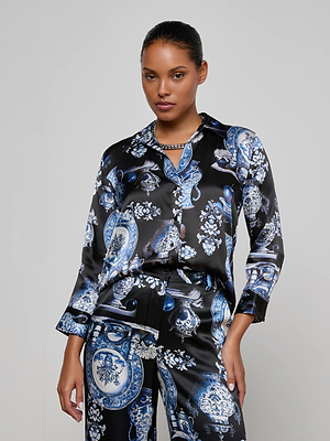 A woman wearing the Dani Silk Blouse by L'AGENCE, featuring a matching baroque-inspired blue and white pattern, stands against a plain light gray background with her hair tied back and a neutral expression.