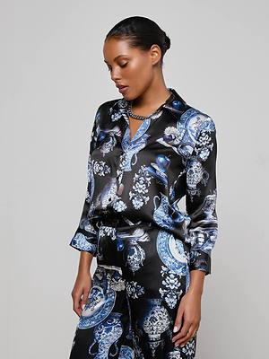 A woman with dark hair in a neat bun wears the Dani Silk Blouse by L'AGENCE, featuring blue and white ornate patterns, standing against a plain light background and looking down thoughtfully.