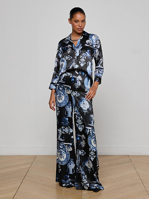 A woman stands indoors wearing the L'AGENCE Dani Silk Blouse, featuring a blue and white ornate pattern on black silk charmeuse, paired with matching wide-leg pajama pants as she poses against a plain, light-colored wall.