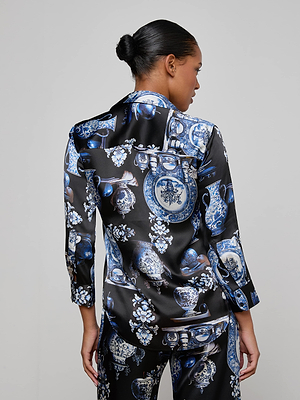 A person with dark hair in a bun is seen from behind, wearing the L'AGENCE Dani Silk Blouse featuring blue and white porcelain vase and floral patterns. The background is plain, light gray.