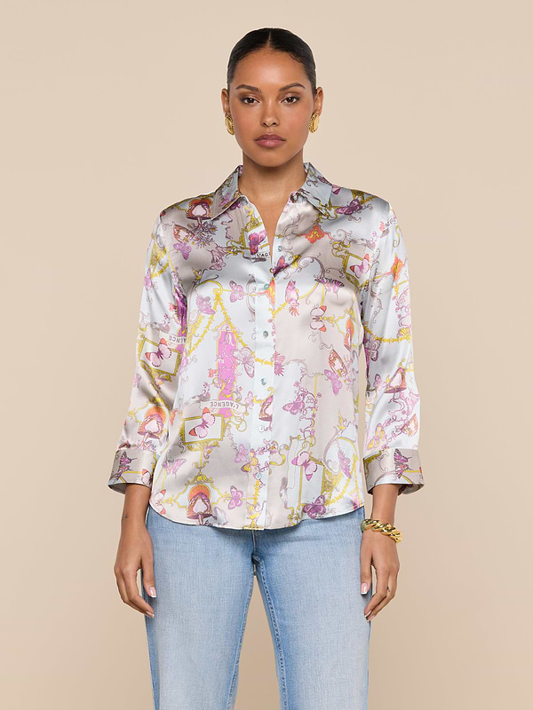 A woman stands against a beige background wearing the Dani Silk Blouse, featuring shiny silk fabric with pink, yellow, and purple designs, paired with light blue jeans. Her hair is pulled back, and she accessorizes with gold earrings and a bracelet.