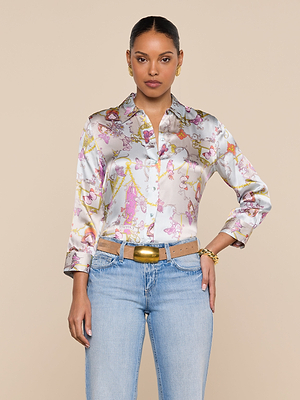 A woman faces forward in the Dani Silk Blouse—shiny with pink and gold designs—paired with light blue jeans, a beige belt, and gold jewelry, set against a plain beige background.