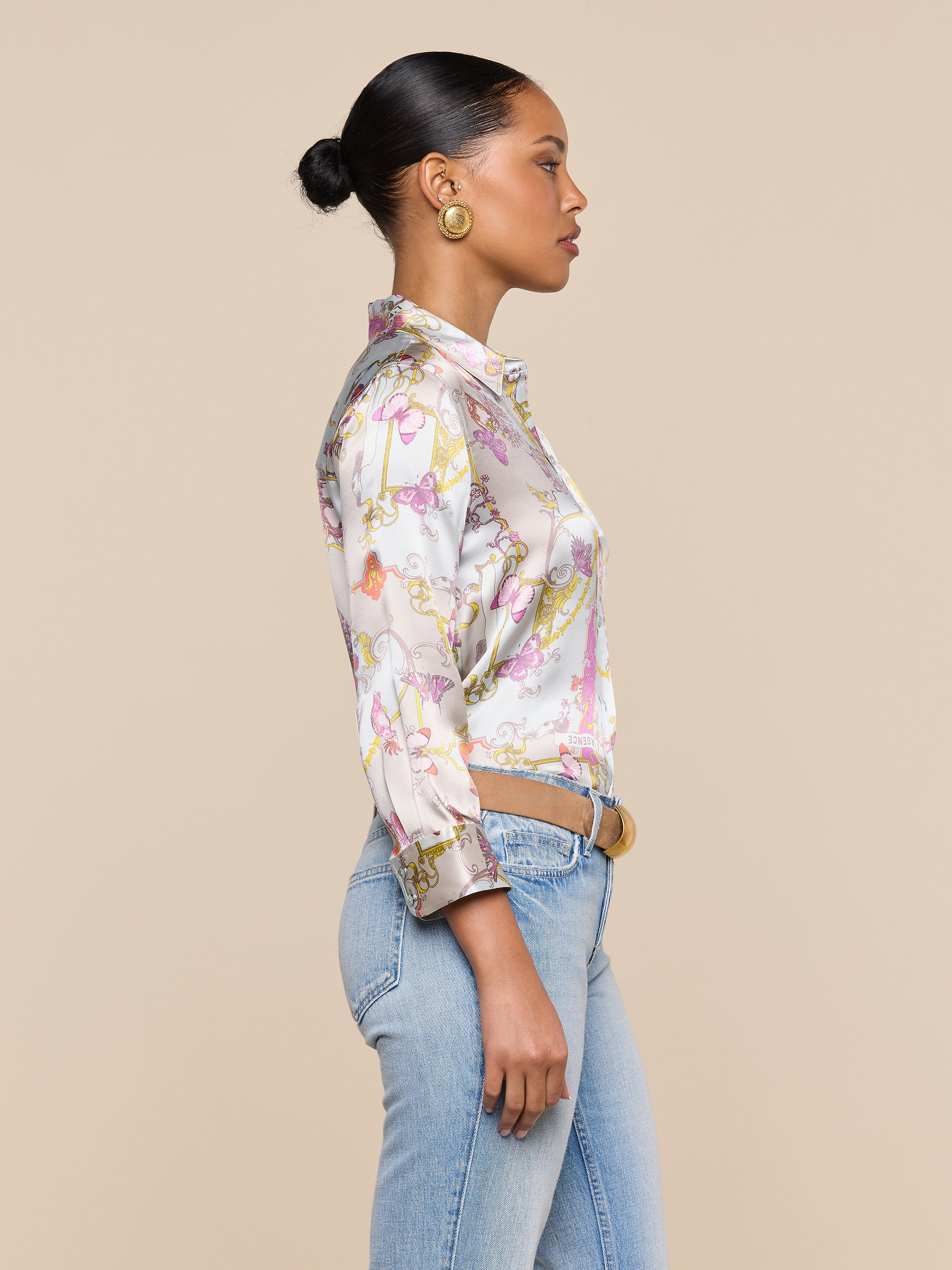 A woman stands in profile against a plain background, wearing a colorful Dani Silk Blouse in a baroque toile print, light blue jeans, gold hoop earrings, and her hair styled in a neat bun.