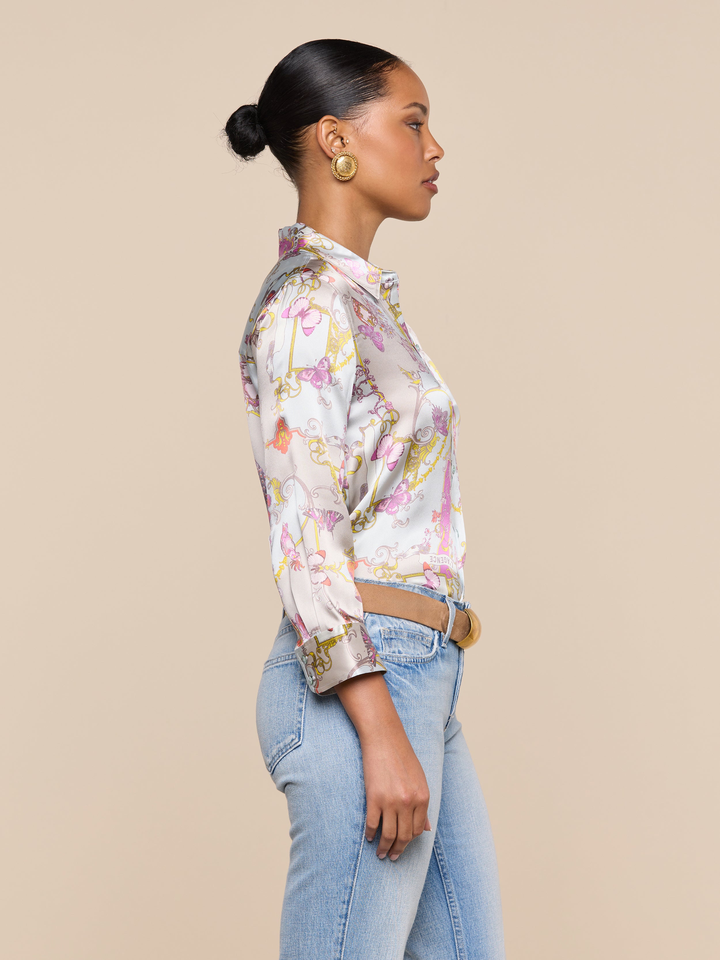 A woman stands in profile against a plain background, wearing a colorful Dani Silk Blouse in a baroque toile print, light blue jeans, gold hoop earrings, and her hair styled in a neat bun.