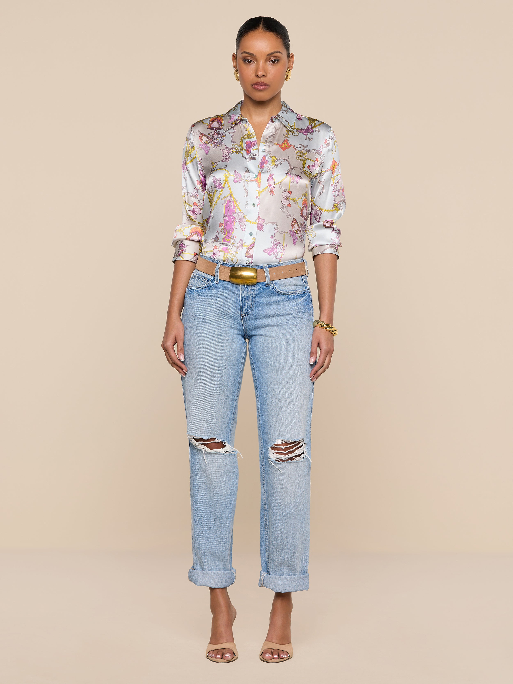 A woman stands against a plain white background, wearing the Dani Silk Blouse in a pastel baroque toile print, paired with light blue ripped jeans, a brown belt, and heeled sandals. Her hair is pulled back and she is facing forward.