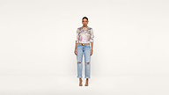 A woman stands against a plain white background, wearing a colorful floral blouse, light blue ripped jeans, a brown belt, and beige heels, looking directly at the camera with arms relaxed by her sides.