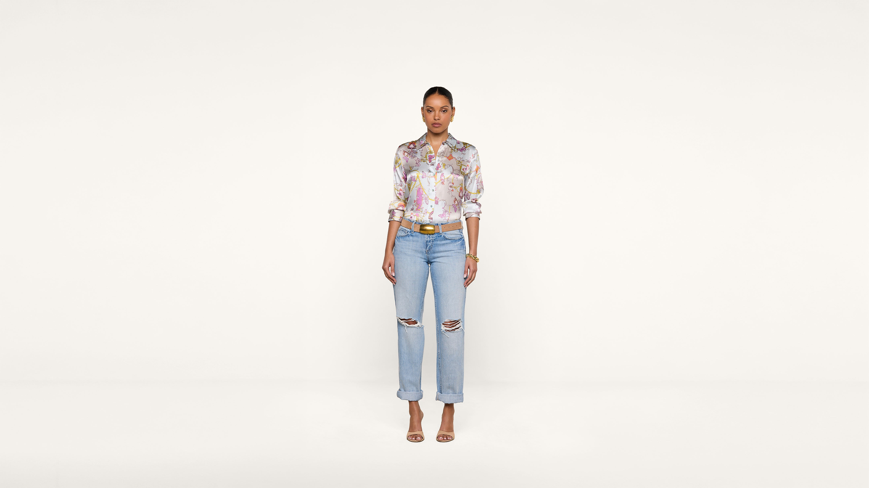A woman stands against a plain white background, wearing a colorful floral blouse, light blue ripped jeans, a brown belt, and beige heels, looking directly at the camera with arms relaxed by her sides.