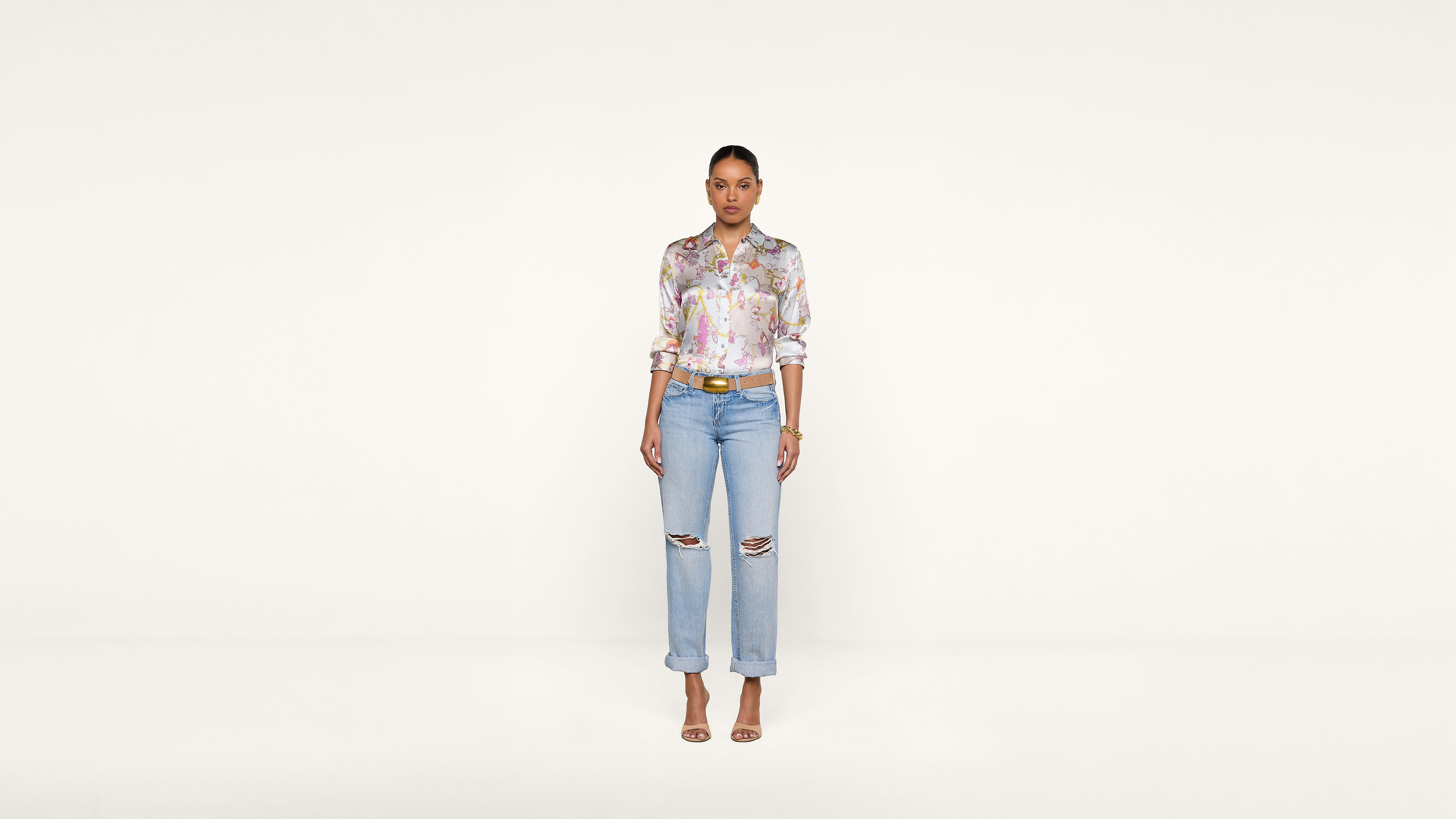A woman stands against a plain white background, wearing a colorful floral blouse, light blue ripped jeans, a brown belt, and beige heels, looking directly at the camera with arms relaxed by her sides.