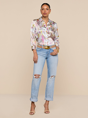 A woman stands against a beige background wearing the Dani Silk Blouse, light blue ripped jeans, a gold belt, and beige heeled sandals. She has her hands in her pockets and accessorizes with earrings and bracelets.