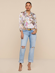A woman stands against a plain white background, confidently posing with her hands in her pockets. She wears the Dani Silk Blouse in a baroque toile print, styled with light blue ripped jeans, a tan belt, and open-toe heels. Her hair is tied back for a polished yet relaxed look.