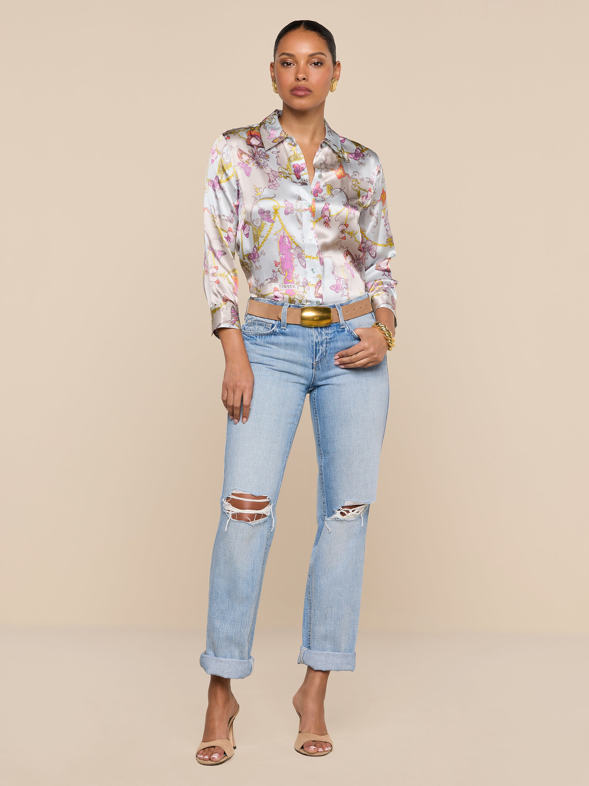 A woman stands against a plain white background, confidently posing with her hands in her pockets. She wears the Dani Silk Blouse in a baroque toile print, styled with light blue ripped jeans, a tan belt, and open-toe heels. Her hair is tied back for a polished yet relaxed look.