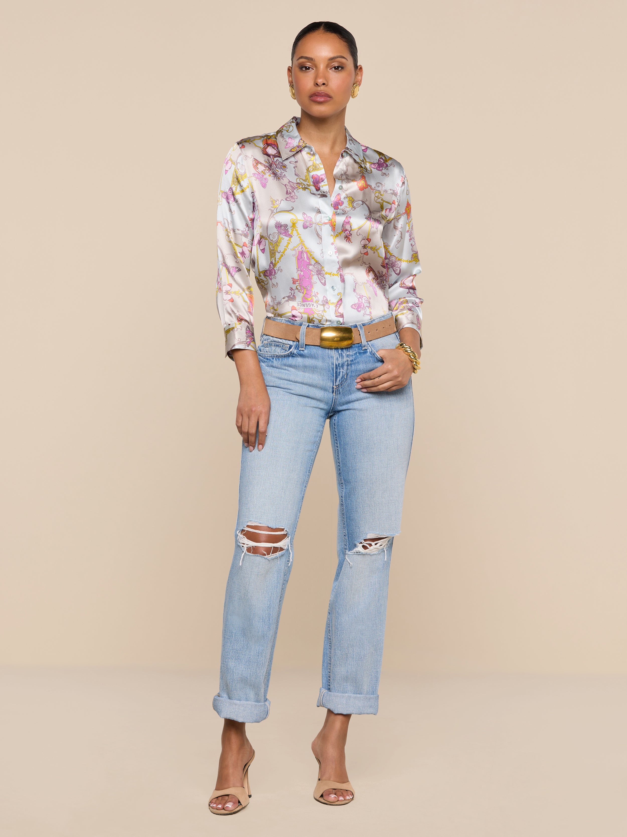 A woman stands against a plain white background, confidently posing with her hands in her pockets. She wears the Dani Silk Blouse in a baroque toile print, styled with light blue ripped jeans, a tan belt, and open-toe heels. Her hair is tied back for a polished yet relaxed look.