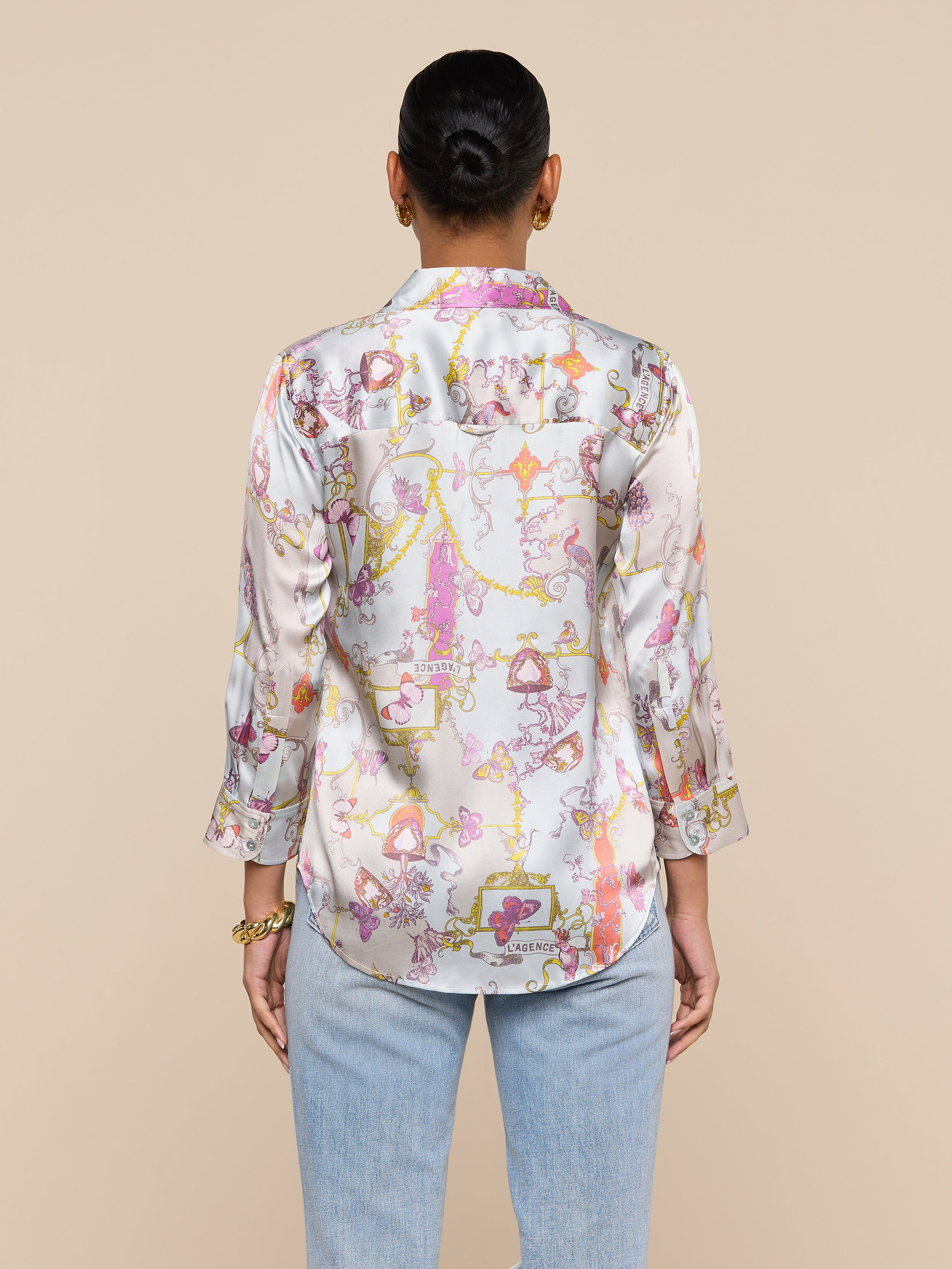 A person with short hair, wearing the Dani Silk Blouse in a pastel pattern with three-quarter sleeves and light blue jeans, is seen from the back against a plain off-white background.