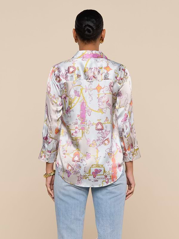 A person stands facing away, wearing the Dani Silk Blouse—a shiny, whimsical top with a baroque toile print—paired with light blue jeans and a gold bracelet. Their hair is tied back, set against a plain beige background.