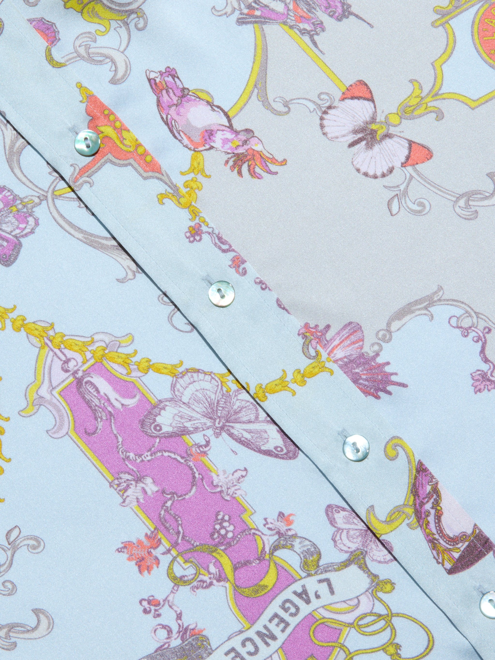 Close-up of the Dani Silk Blouse, a light blue patterned silk button-down shirt with shiny buttons, adorned with pink, purple, and yellow ornate designs featuring butterflies and decorative baroque toile print motifs.