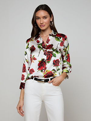 A woman with long brown hair wears the Dani Silk Blouse with a red rose print, paired with white pants, a black belt, and hoop earrings, standing against a plain light background.
