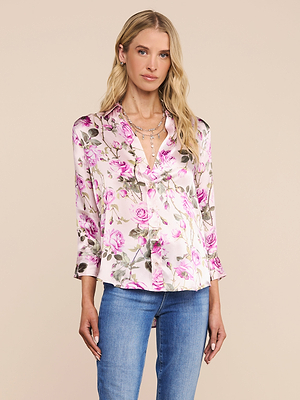 A woman with long blonde hair wears the Dani Silk Blouse, featuring a light pink rose print and deep V-neck, paired with blue jeans, standing against a plain beige background.
