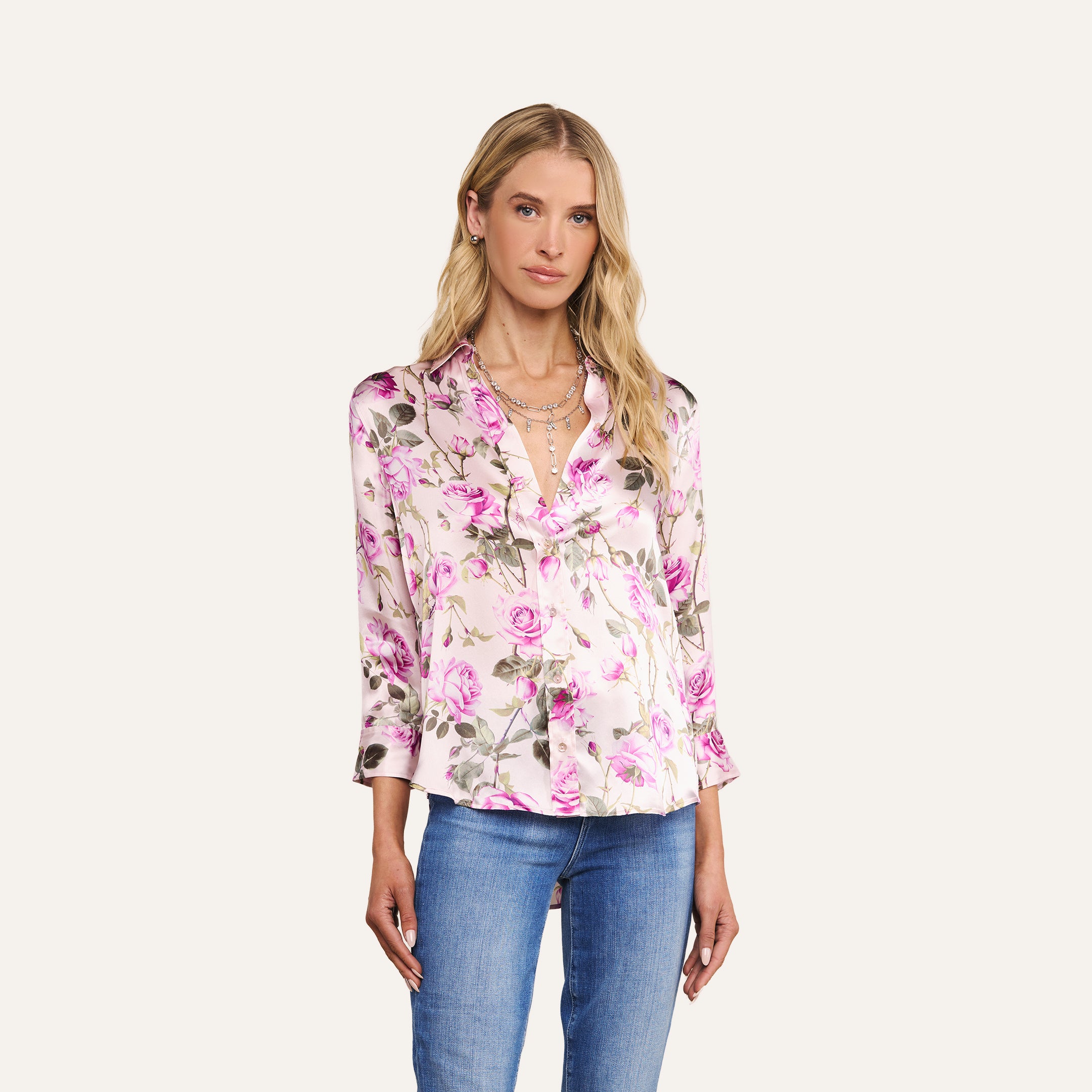 A woman wears the Dani Silk Blouse, a button-down top adorned with a pink rose print, paired with blue jeans and standing against a plain, light background.