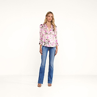A woman with blonde hair stands against a white background, wearing a pink rose print Dani Silk Blouse, blue jeans, and beige heeled sandals. She has a neutral expression with her arms relaxed at her sides.
