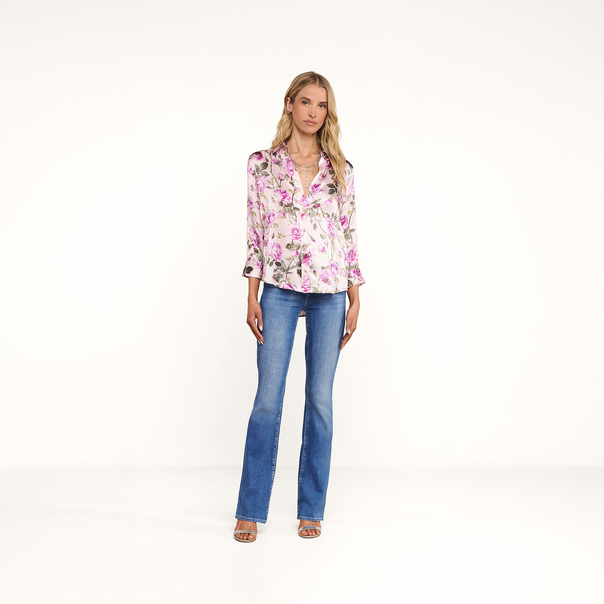 A woman with blonde hair stands against a white background, wearing a pink rose print Dani Silk Blouse, blue jeans, and beige heeled sandals. She has a neutral expression with her arms relaxed at her sides.