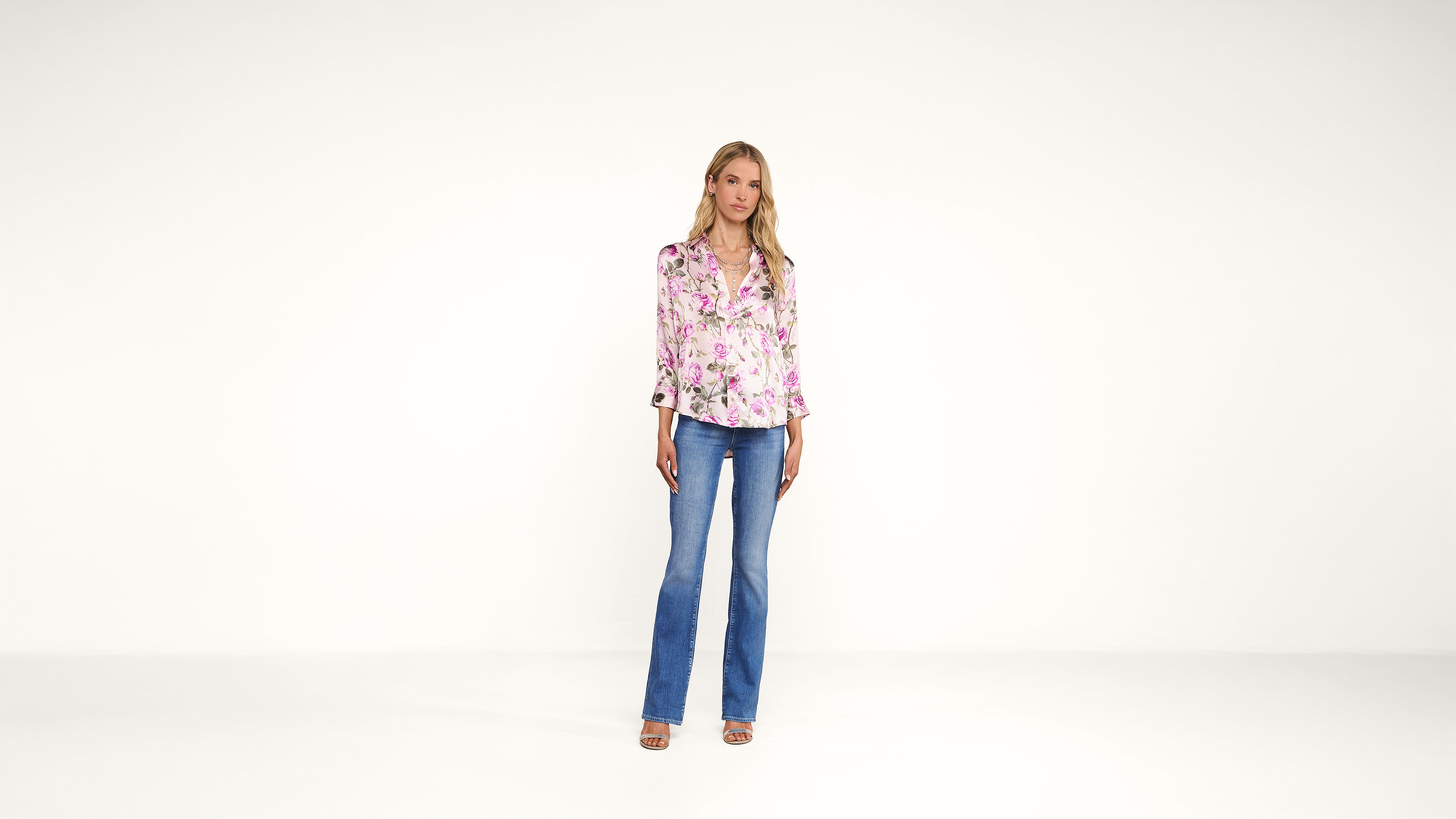 A woman stands against a plain white background, wearing a pink floral blouse, blue flared jeans, and sandals. She has blonde hair and a neutral expression, with her arms relaxed by her sides.