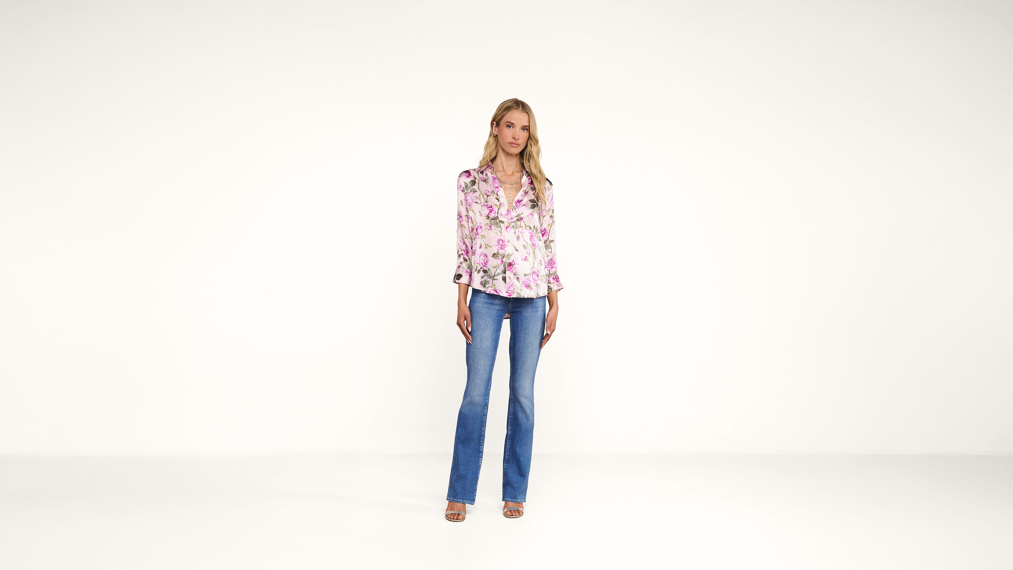A woman stands against a plain white background, wearing a pink floral blouse, blue flared jeans, and sandals. She has blonde hair and a neutral expression, with her arms relaxed by her sides.