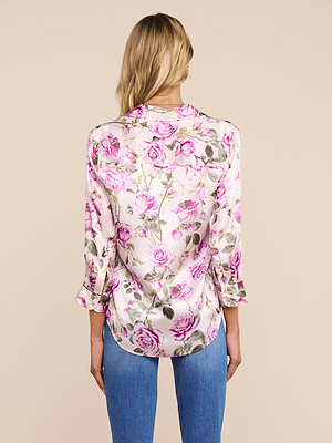 A woman with long blonde hair, seen from behind, wears the Dani Silk Blouse in light pink with a rose print and blue jeans, standing against a beige background.