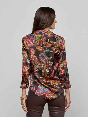 A woman with long brown hair, seen from the back, wears the L'AGENCE Dani Silk Blouse in a colorful paisley print and brown leather pants, standing against a plain light gray background.
