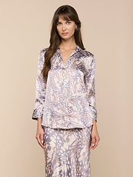 Una donna dai lunghi capelli castani modella la blusa di seta Dani e la gonna abbinata in morbide tonalità pastello con stampa di ali di farfalla, in piedi su un semplice sfondo beige e rivolta in avanti con unespressione neutra.